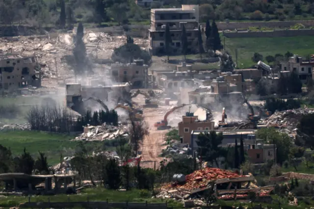 In a picture taken from far away, piles of rubble can be seen amongst a few houses that remain standing. Dust rises into the air as half a dozen yellow excavators look to be demolishing buildings