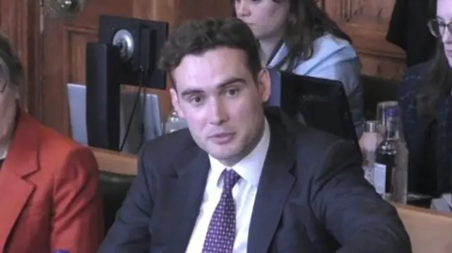 Henry Tufnell sits at a select committee table, dressed in a suit and purple tie.