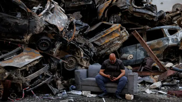 A man sits beside piled damaged cars at the site of an Israeli strike in Beirut