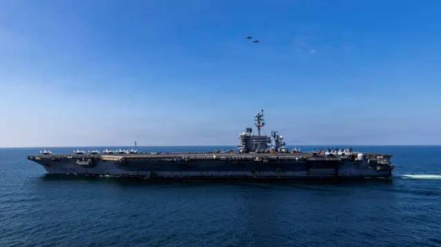 A photo of the aircraft carrier USS Abraham Lincoln sailing, two fighter jets are flying above it and several can be seen parked on its deck