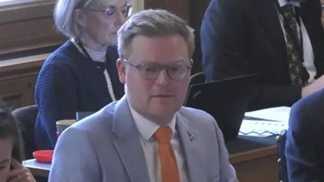 A man with short hair, glasses and a light blue suit with an orange tie.