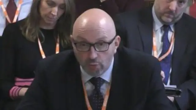 A man in a dark black suit. He is bald and wearing an orange lanyard.