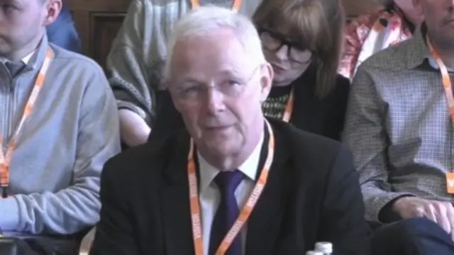 Chris Train speaks as he sits at a parliamentary table, wearing a suit and tie and orange lanyard.
