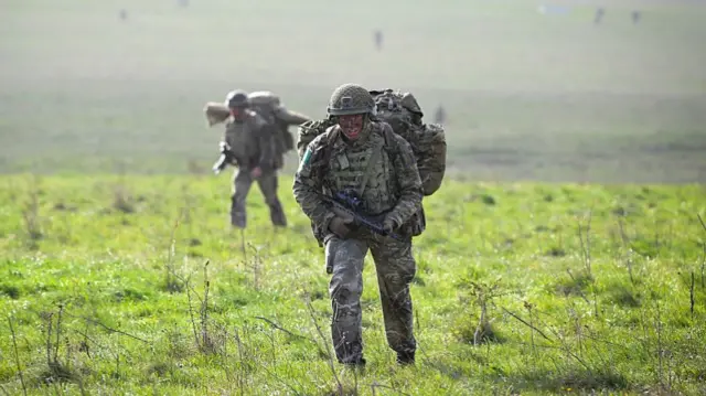 Two soldiers in a field walking towards the camera. They wear the uniform, a large backpack and hold large guns