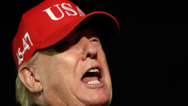 Donald Trump speaks to the members of the media. He wears a red 'USA' cap