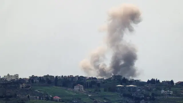 Smoke rises from several buildings on top of a hill, with green grasses and trees also visible. The sky above the horizon is grey.