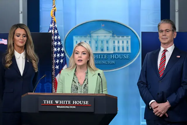 Three people in suits stand in front of a blue backdrop with the White House logo