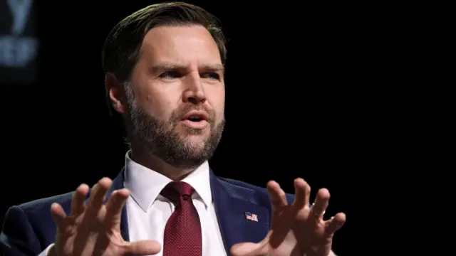 JD Vance on stage with his hands clawed expressively while talking and dressed in a navy suit with a red tie.