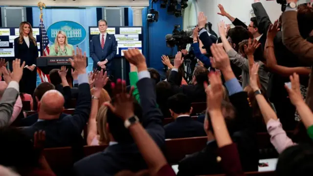 A large group of members of the press raise their hands to ask questions in the White House briefing room