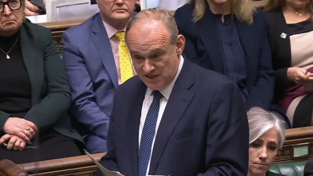 Ed Davey in dark suit stands reading from papers in parliamentary chamber