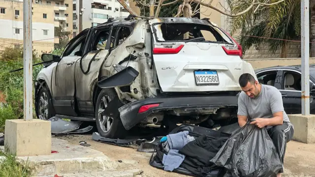 A man sat next to a car which has been burnt, he is placing items into a bin bag in Tyre, southern Lebanon.