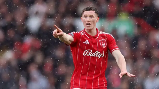 Elliot Anderson, wearing a red Nottingham Forest shirt with white pinstripes, points with his right arm with his mouth open to shout
