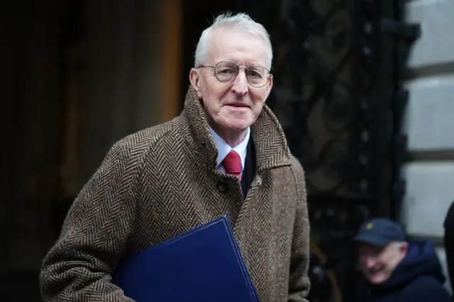 a man in a tweed coat looks at the camera. He is wearing a red tie, white shirt and glasses.
