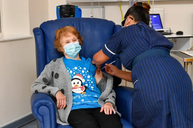 An elderly woman sitting on a blue leather armchair receives a jab from a matron in a striped dark blue dress