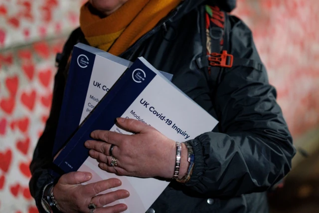 A woman in a green rain coat holds two copies of a document titled UK Covid-19 Inquiry. Behind her, in the blurred background, is part of the Thames embankment covered in red hearts commemorating victims of coronavirus