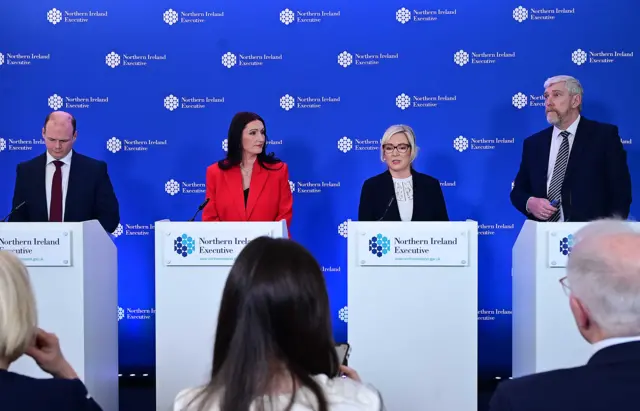 Two women and two men stand in front of white podiums that say 'Northern Ireland Executive'. They are standing in front of a blue background with the same phrase.
