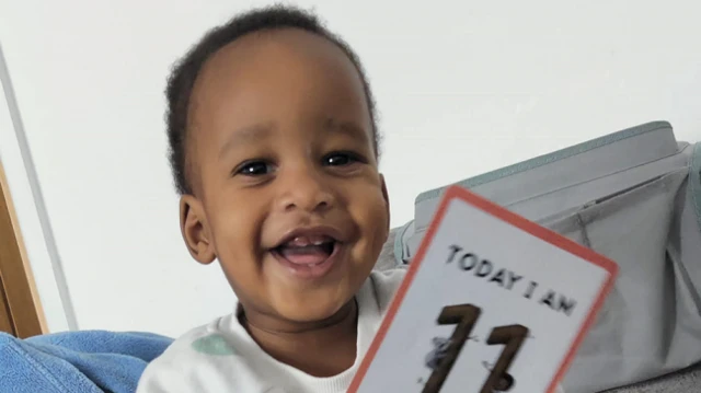 Noah Sibanda has short curly black hair and is smiling. He has a white jumper on and is holding a card that says "today I am 11 months old".