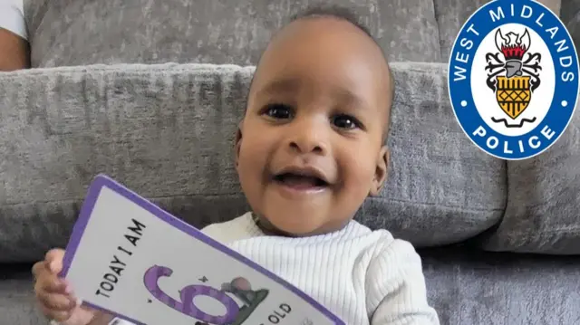 Noah is six months old in the picture, and is wearing a white babygro and holding a card that is purple and white, and says "Today I am six months old". He is sitting against a grey sofa, and has tufts of black hair. He is smiling at the camera
