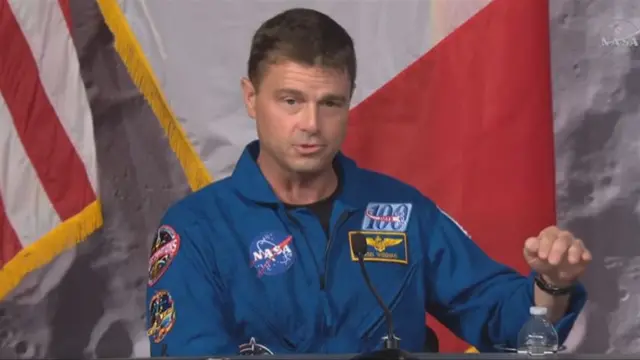 Reid Wiseman speaking in front of a microphone, wearing a blue NASA outfit