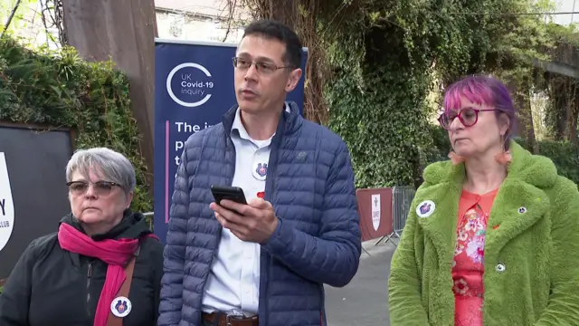 From left to right: A short woman in black coat and fuchsia scarf, a tall man with cropped black air, an open blue puffer jacket holding his phone in his left hand while speaking; a woman with purple hair in a green fur coat. They're standing outside in front of a blue banner reading UK Covid-19 Inquiry in the top right corner