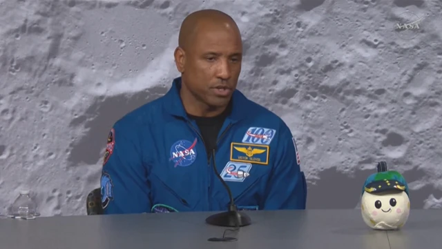 Victor Glover speaking in front of a microphone, wearing a blue NASA outfit