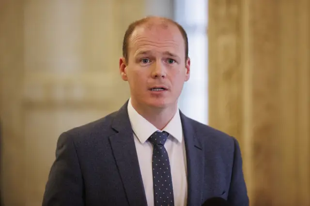 Gordon Lyons stands facing past the camera. He is wearing a navy suit, navy tie with yellow spots and a white shirt underneath.