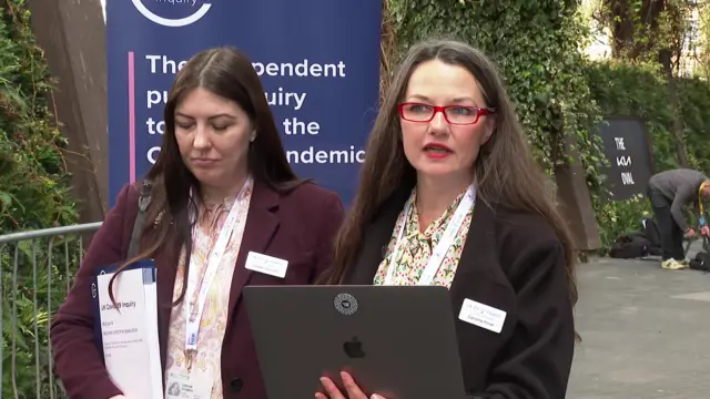 Woman with red glasses wearing a black suit with floral shirt holds an Apple Mac computer while addressing the media outside the headquarters of the Covid inquiry in London. A woman with long brown hair in a plum suit stands next to her, to the right