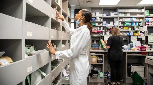 A woman wearing a lab coat and Covid mask browsing the shelf of a pharmacy, to prepare medication to deliver to patients