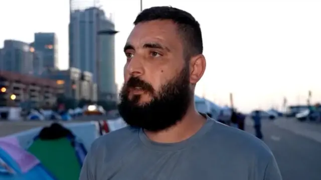 Displaced Lebanese resident speaks to camera for an interview as he stands near a group of tents