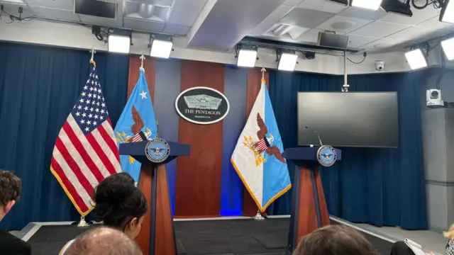 Two lecterns are seen with a US flag and a TV next to Pentagon branding