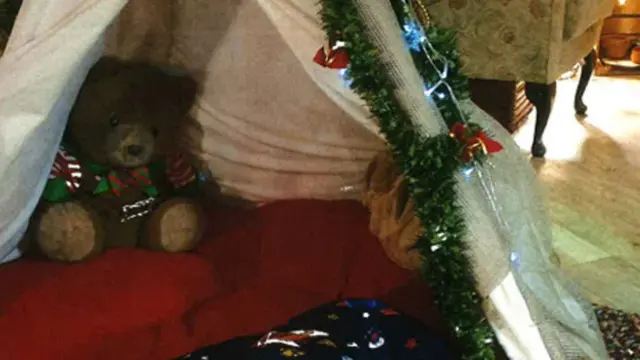 A small teepee style tent for young children. It has white fabric on the outside and a red cushion on the inside with a brown teddy beat in there. There are fairy lights and tinsel wrapped around the outside.