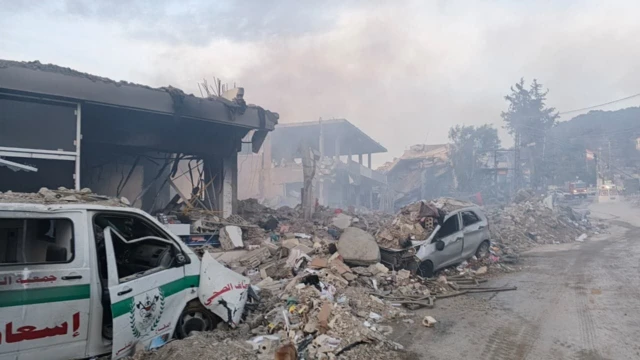 A large amount of rubble by the side of the road, next to a destroyed ambulance and car as well as at least three partially destroyed buildings following strikes