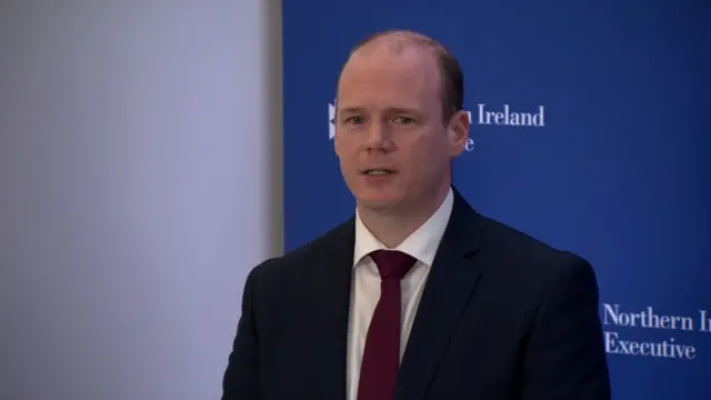 Gordon Lyons stands in front of a blue screen. He is wearing a suit and tie.