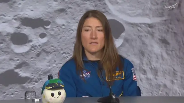 Christina Koch speaking in front of a microphone, wearing a blue NASA outfit