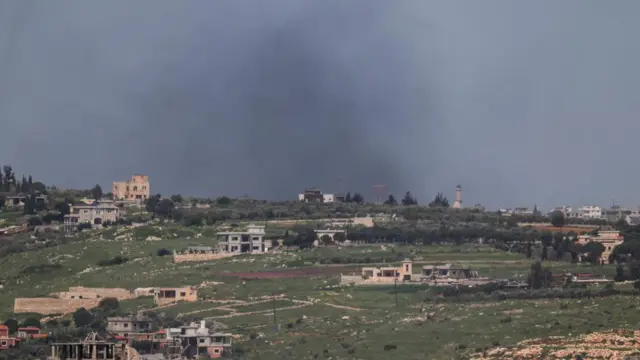 Smoke rises from a settlement on top of a grassy hill. The sky is grey behind the black smoke rising from the horizon.