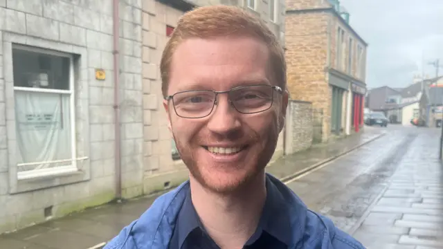 Ross Greer, a man with ginger hair and glasses, smiling while standing on a cobbled street.