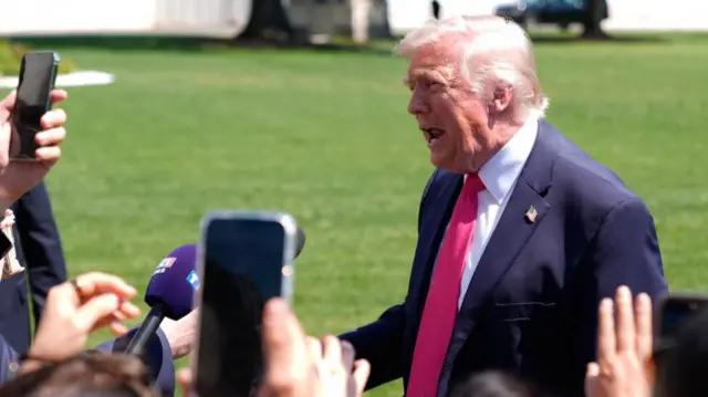 President Donald Trump addressing the press on the south lawn of the White House. He's in a dark blue suit, white shirt and red tie, in front of him several mics and phones held up by reporters