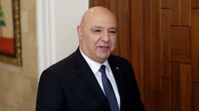 Joseph Aoun, a bald man wearing a dark suit, white shirt and blue tie, smiles off-camera as he walks down a corridor.