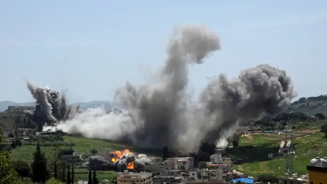 Large plumes of smoke rise immediately after strike impact on several buildings in a green valley, with flames visible in the foreground.