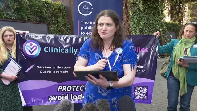 A woman in a blue dress holds an iPad while holding a small mic as she addresses reporters. Behind her are two women, one to her right and one to her left, holding a banner for Clinically Vulnerable Families
