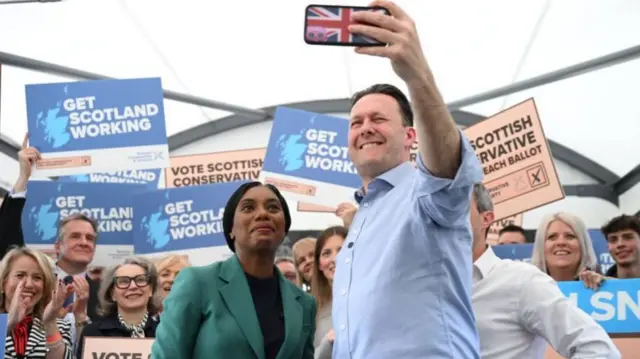 Kemi Badenoch wearing a green jacket, standing next to Russell Findlay in a blue shirt, he has his arm outstretched holding an iphone in a union jack case.