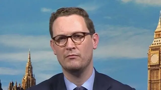 Headshot of Jones who wears glasses and sits in front of a background which shows the skyline of London including a part of Big Ben