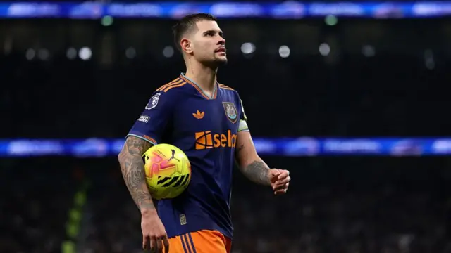 Bruno Guimaraes dressed in a navy top with bright orange detailing and bright orange shorts. He has a yellow football tucked under his arm