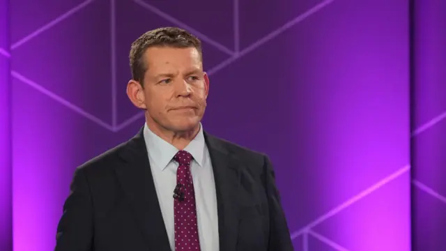 Rhun ap Iorwerth, a man with short brown hair wearing a black suit jacket, white shirt and purple spotted tie
