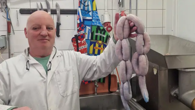 A butcher holding up blue skinned-sausages inside a butchers back room