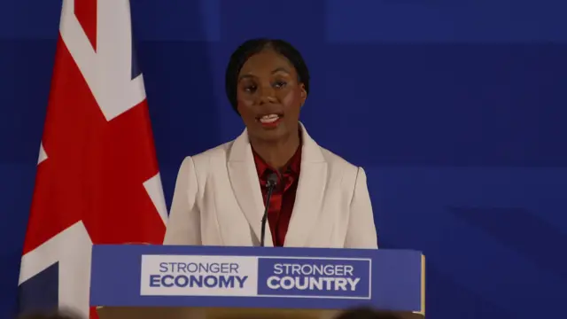 Kemi Badenoch holding a pres conference, she is standing in front of a blue background and a Union Jack flag