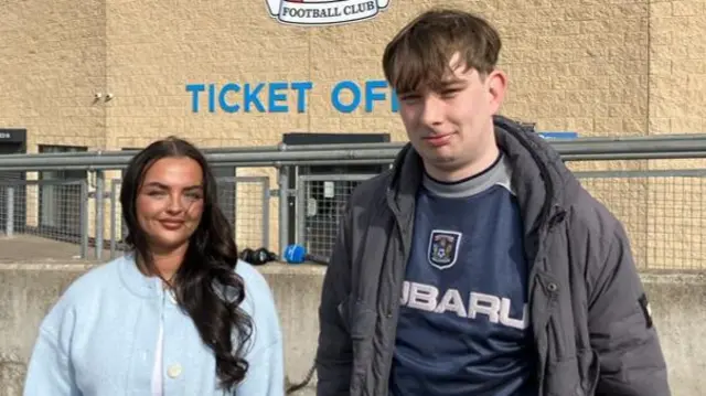 Coventry City fans Demi-Rae on the left and Alex on her right. Demi-Rae is wearing a sky blue cardigan and has long dark hair. Alex is wearing a navy blue Coventry shirt with a black coat. They're stood in front of the football ground with the Ticket Office sign on the wall behind them.