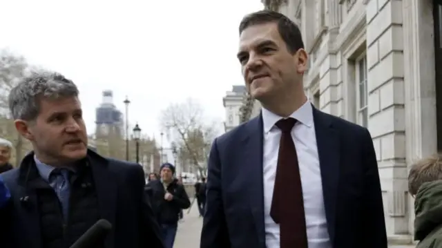 Olly Robbins is approached by a journalist on a central London street. He wears a dark blue suit, white shirt and brown tie. He has short brown hair and is smiling.