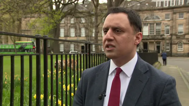 Head shot of Sarwar standing on a street next to a park with grass and daffodils as he speaks