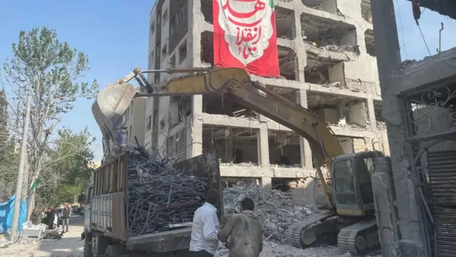 A digger in front of a destroyed building with a flag hanging off it